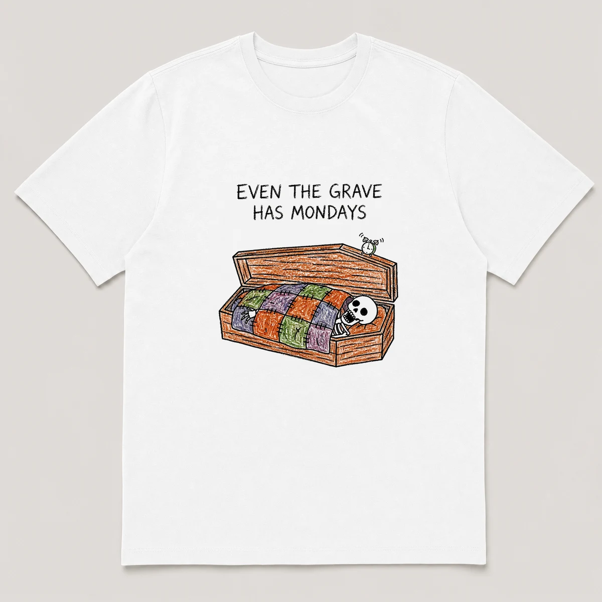 Front view of a White Tee printed with a skeleton yawns under a patchwork quilt inside an open coffin while a tiny alarm clock rings on the lid and the caption EVEN THE GRAVE HAS MONDAYS, White front view