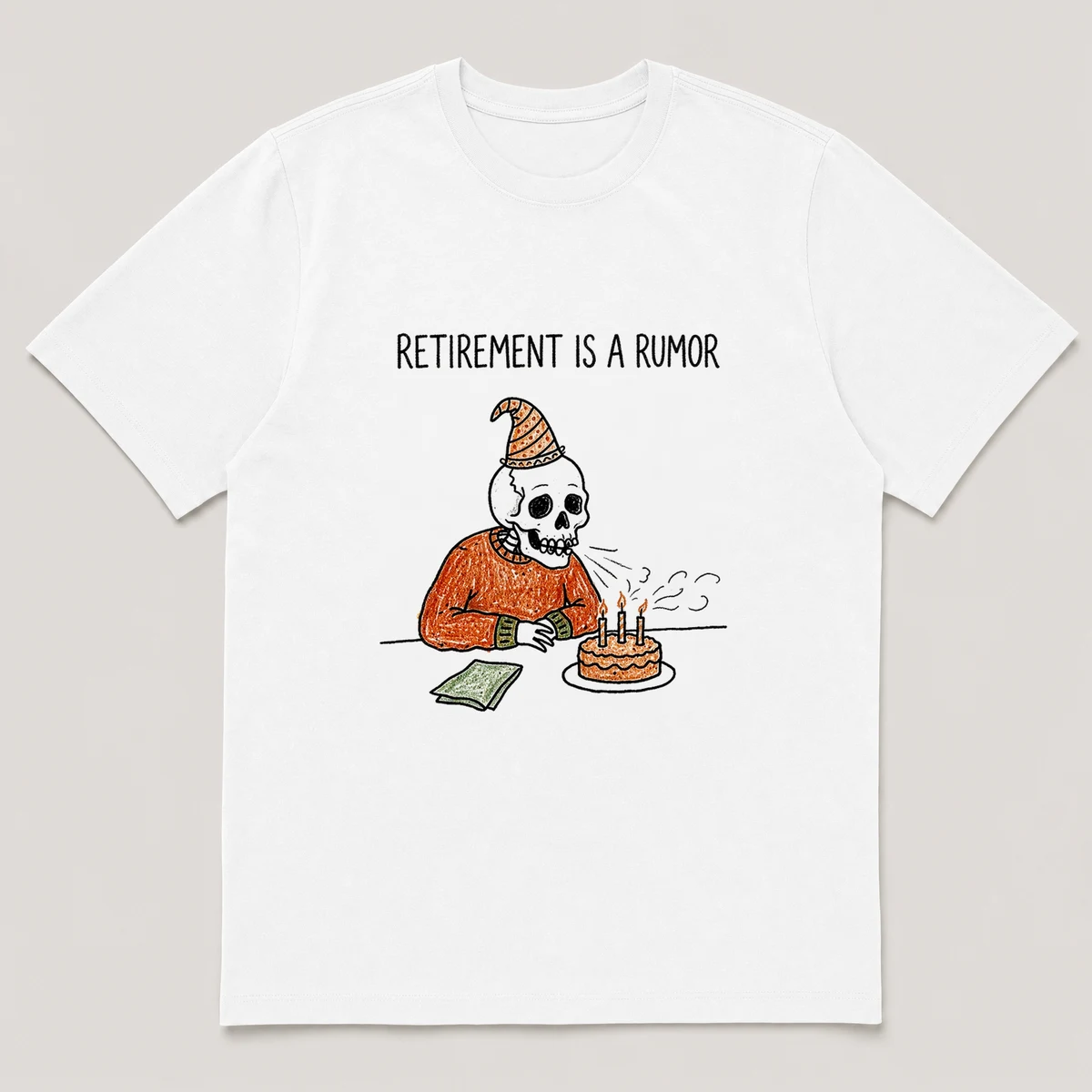 Front view of a White Tee printed with old skeleton in a party hat blows out candles on a small cake at a desk and the caption RETIREMENT IS A RUMOR, White front view